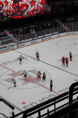 A hockey game is taking place on an indoor ice rink. Players, dressed in red and white uniforms, are gathered around the center ice area, marked with a large star logo. Several referees in black and white striped uniforms are also on the ice. Above the rink is a large digital scoreboard displaying red star graphics. The stands are partially filled with spectators.