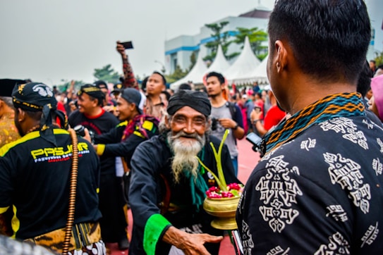 A festive outdoor gathering with a focus on cultural attire and traditional activities. Several individuals are dressed in colorful and patterned clothing, engaging in a celebration. One person is prominently holding a decorative container with flowers and grass. A diverse group of people seems to be enjoying the event.