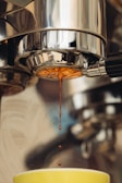 A close-up of a coffee machine with espresso dripping from the portafilter into a cup below. The focus highlights the flow of the coffee and the stainless steel surface of the machine reflects light, creating a polished look.