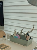 An organized desk setup includes a white table surface with a computer monitor partially visible on the left. A light wooden chair is seen, and next to it, a transparent cup with 'BAES' written on it. Various stationary items such as notebooks and papers in different colors are neatly arranged in a green Vitra organizer. Decorative items like dried eucalyptus branches are placed in the organizer along with some skincare bottles.