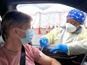 A healthcare worker wearing a colorful headscarf, mask, and protective gloves administers a vaccine to a man sitting in a car. The setting appears to be a drive-through vaccination site with white tents and orange traffic cones visible in the background. Both individuals are wearing face masks, and the health worker is dressed in a light-colored protective gown.