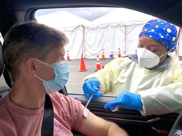 A healthcare worker wearing a colorful headscarf, mask, and protective gloves administers a vaccine to a man sitting in a car. The setting appears to be a drive-through vaccination site with white tents and orange traffic cones visible in the background. Both individuals are wearing face masks, and the health worker is dressed in a light-colored protective gown.