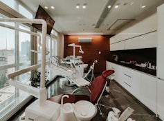 A modern dental clinic interior with dental chairs lined up near large windows, bright overhead lighting, and cabinetry. The room is well-organized and appears clean and professional, with a neutral color scheme and urban city view through the windows.