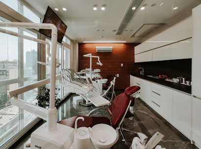 A modern dental clinic interior with dental chairs lined up near large windows, bright overhead lighting, and cabinetry. The room is well-organized and appears clean and professional, with a neutral color scheme and urban city view through the windows.