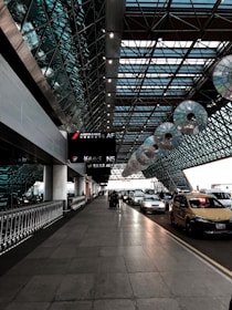 The image depicts an airport taxi drop-off area under a modern, expansive glass and steel canopy. A line of taxis and cars is parked on the right side, while the left features a row of luggage carts. Overhead, signage for airlines is visible, and the architecture has a geometric, futuristic design with patterns created by the glass panels.