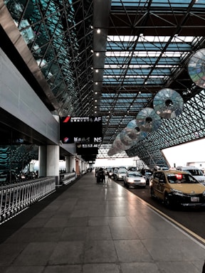 The image depicts an airport taxi drop-off area under a modern, expansive glass and steel canopy. A line of taxis and cars is parked on the right side, while the left features a row of luggage carts. Overhead, signage for airlines is visible, and the architecture has a geometric, futuristic design with patterns created by the glass panels.