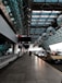 The image depicts an airport taxi drop-off area under a modern, expansive glass and steel canopy. A line of taxis and cars is parked on the right side, while the left features a row of luggage carts. Overhead, signage for airlines is visible, and the architecture has a geometric, futuristic design with patterns created by the glass panels.