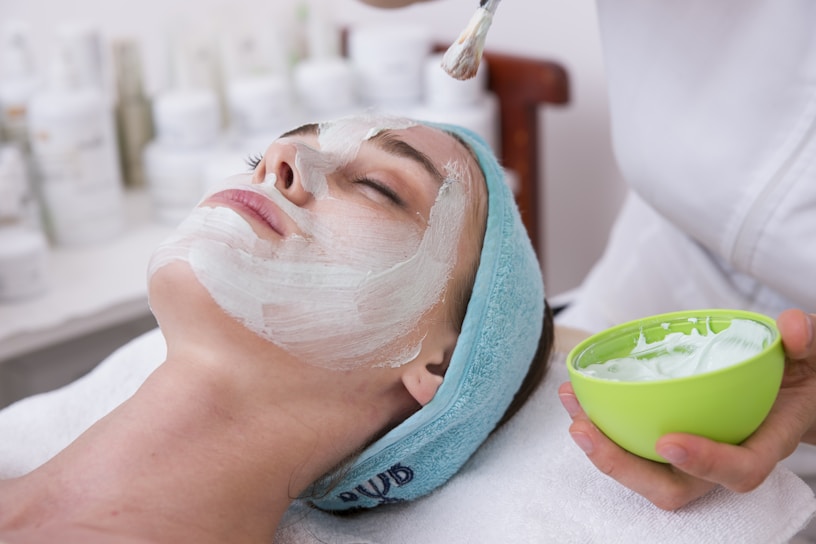 A person is lying down with a white facial cream applied to the face. A hand is holding a green bowl containing more cream, using a brush to apply it. The individual appears relaxed and is wearing a blue headband with their eyes closed. The background shows blurred bottles, likely skincare products.