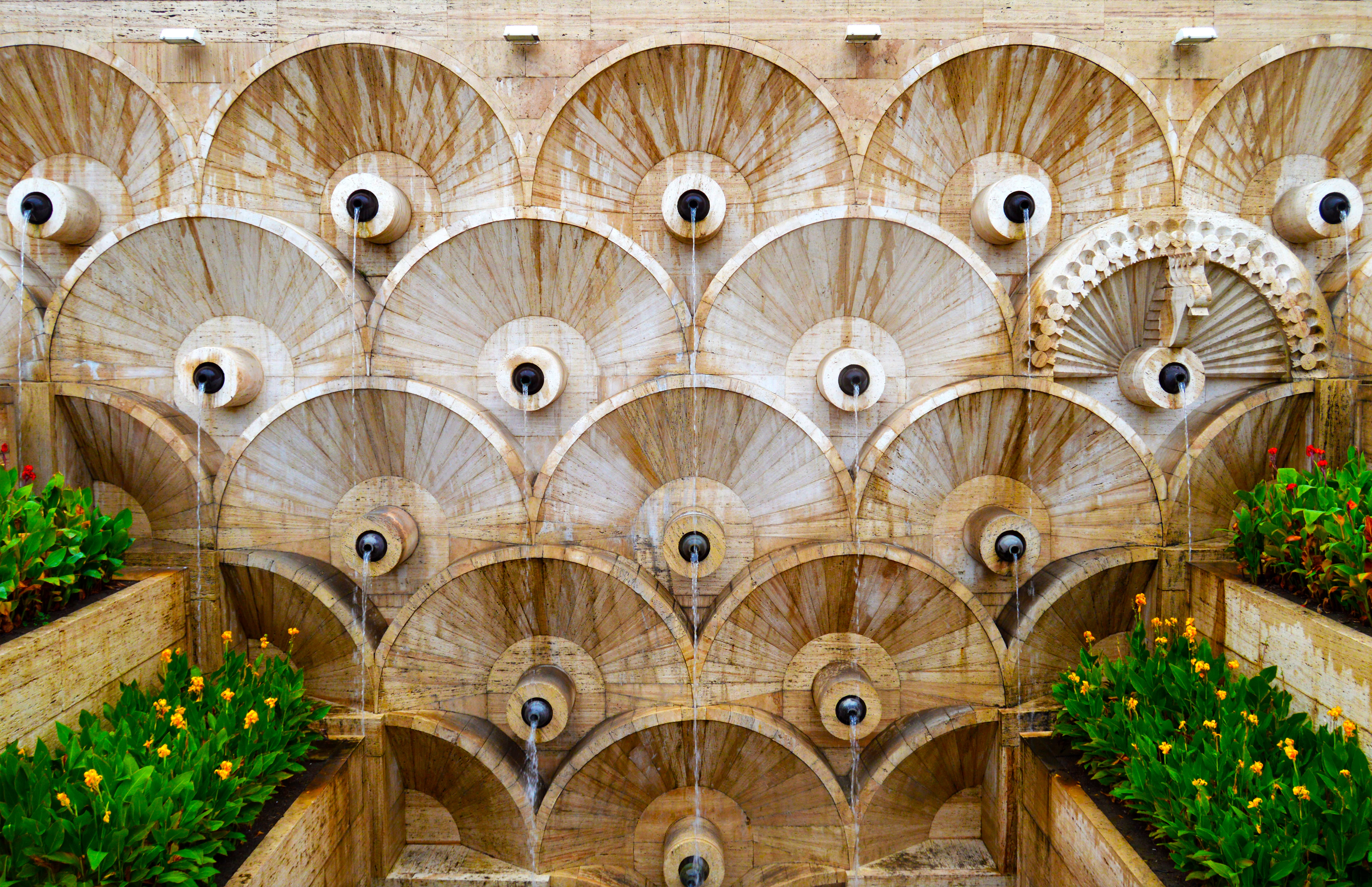 The tiered stone fountain wall of the Cascade complex in Yerevan, its repeated fan and shell carvings flanked by planted beds of flowers.