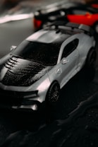 A sleek, silver sports car with black accents and visible raindrops on its surface is prominently featured. Out of focus in the background are two additional cars, one black and the other red. The setting appears to be wet, perhaps suggesting a rainy environment.
