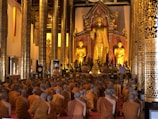 A large group of monks clad in orange robes are seated on the floor in a grand temple hall, facing a magnificent large golden Buddha statue. The hall is adorned with ornate, gilded columns and intricate decorative patterns.
