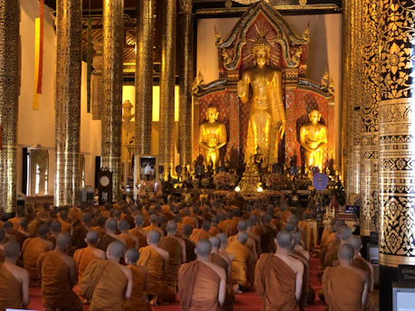 A large group of monks clad in orange robes are seated on the floor in a grand temple hall, facing a magnificent large golden Buddha statue. The hall is adorned with ornate, gilded columns and intricate decorative patterns.