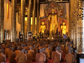 A large group of monks clad in orange robes are seated on the floor in a grand temple hall, facing a magnificent large golden Buddha statue. The hall is adorned with ornate, gilded columns and intricate decorative patterns.