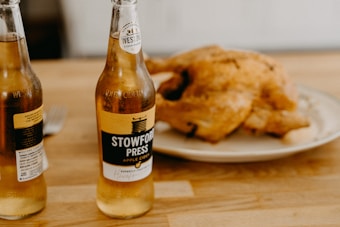 Two bottles of apple cider are placed on a wooden table alongside a whole roasted chicken on a white plate. The labels on the bottles read 'Stowford Press Apple Cider,' and there is a fork visible in the background.