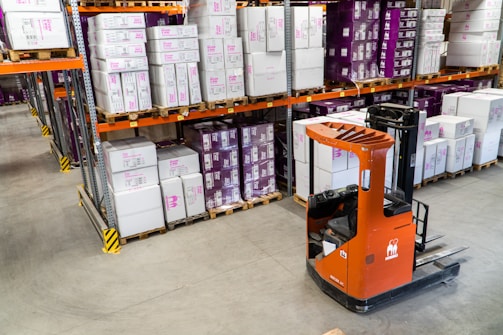 A warehouse interior featuring shelves stacked with cardboard boxes and packages, all organized neatly on wooden pallets. An orange forklift is parked on the concrete floor, illustrating a typical industrial storage scene.