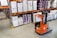 A warehouse interior featuring shelves stacked with cardboard boxes and packages, all organized neatly on wooden pallets. An orange forklift is parked on the concrete floor, illustrating a typical industrial storage scene.