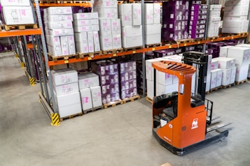 A warehouse interior featuring shelves stacked with cardboard boxes and packages, all organized neatly on wooden pallets. An orange forklift is parked on the concrete floor, illustrating a typical industrial storage scene.