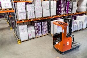 A warehouse interior featuring shelves stacked with cardboard boxes and packages, all organized neatly on wooden pallets. An orange forklift is parked on the concrete floor, illustrating a typical industrial storage scene.