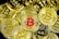 A collection of cryptocurrency coins with the Bitcoin symbol prominently displayed, most of which are gold, except for one coin in the center that features a red Bitcoin symbol.