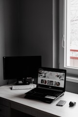A minimalist workspace features a laptop with an open website displaying food images, accompanied by a wireless keyboard and mouse, set against a dark monitor and a window letting in natural light.