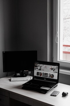 A minimalist workspace features a laptop with an open website displaying food images, accompanied by a wireless keyboard and mouse, set against a dark monitor and a window letting in natural light.