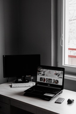 A minimalist workspace features a laptop with an open website displaying food images, accompanied by a wireless keyboard and mouse, set against a dark monitor and a window letting in natural light.