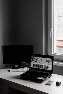 A minimalist workspace features a laptop with an open website displaying food images, accompanied by a wireless keyboard and mouse, set against a dark monitor and a window letting in natural light.