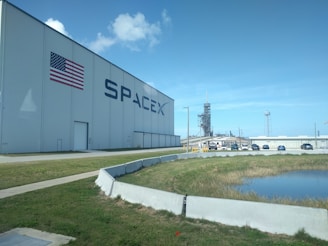 A large warehouse-like building with the American flag and the SpaceX logo prominently displayed. In the background, a rocket launch pad structure is visible. Several cars are parked in the vicinity with a water body and grassy area in the foreground. The sky is clear with a few clouds.