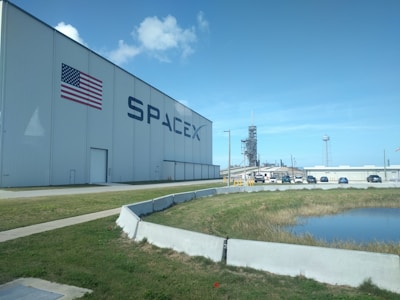 A large warehouse-like building with the American flag and the SpaceX logo prominently displayed. In the background, a rocket launch pad structure is visible. Several cars are parked in the vicinity with a water body and grassy area in the foreground. The sky is clear with a few clouds.