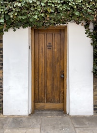 A wooden door with vertical panels, surrounded by white walls and overhung with lush green ivy. The door has a rustic appearance with a round doorknob on the right side. The text 'The Garden Door' is displayed on the door's surface. The bottom of the image shows a stone pavement in front of the door.