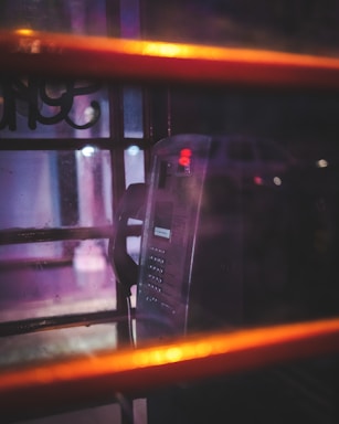 A dimly lit telephone booth with a vintage payphone inside. The booth is seen through glass panels with a warm and moody lighting, highlighting reflections and shadows.
