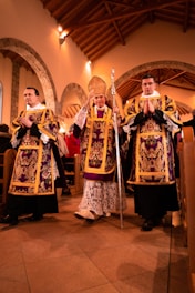 Three religious figures dressed in ornate robes stand in a church setting with wooden beams and stone arches visible in the background. The central figure holds a decorative staff and wears a mitre.