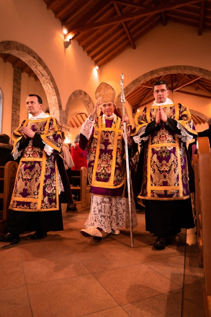 Three religious figures dressed in ornate robes stand in a church setting with wooden beams and stone arches visible in the background. The central figure holds a decorative staff and wears a mitre.