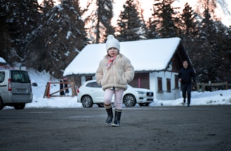 A young child wearing a furry coat, beanie, and boots stands on a snowy road. In the background, there is a snow-covered house and several parked cars. The scene is set in a winter landscape with trees covered in snow.