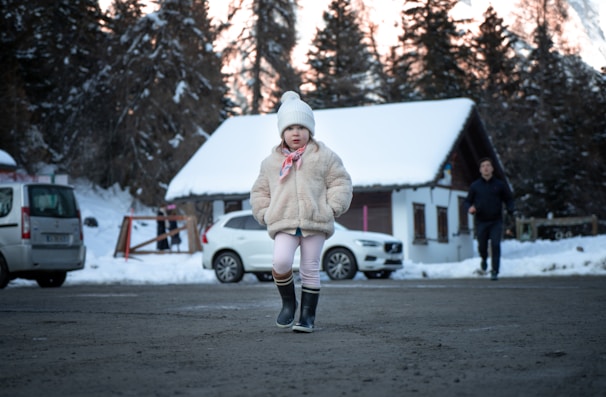 A young child wearing a furry coat, beanie, and boots stands on a snowy road. In the background, there is a snow-covered house and several parked cars. The scene is set in a winter landscape with trees covered in snow.