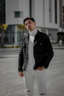A person wearing a black jacket over a white turtleneck sweater stands confidently in an urban environment. The outfit is complemented by ripped white jeans and a watch on the left wrist. The background features modern architecture with large glass windows and bright sunlight casting shadows.