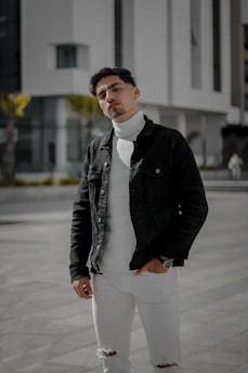 A person wearing a black jacket over a white turtleneck sweater stands confidently in an urban environment. The outfit is complemented by ripped white jeans and a watch on the left wrist. The background features modern architecture with large glass windows and bright sunlight casting shadows.