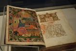 An open illuminated manuscript with a richly decorated image on the left page, depicting a figure in robes sitting with a writing instrument, surrounded by ornate architectural elements and greenery. The right page features text written in a decorative script.