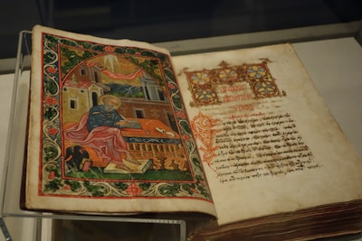 An open illuminated manuscript with a richly decorated image on the left page, depicting a figure in robes sitting with a writing instrument, surrounded by ornate architectural elements and greenery. The right page features text written in a decorative script.