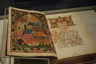 An open illuminated manuscript with a richly decorated image on the left page, depicting a figure in robes sitting with a writing instrument, surrounded by ornate architectural elements and greenery. The right page features text written in a decorative script.