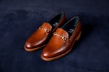 A pair of elegant brown leather loafers with a sleek design and gold detailing on a dark, textured background. The shoes have a polished finish and visible stitching around the edges.