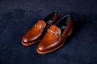 A pair of elegant brown leather loafers with a sleek design and gold detailing on a dark, textured background. The shoes have a polished finish and visible stitching around the edges.