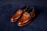 A pair of elegant brown leather loafers with a sleek design and gold detailing on a dark, textured background. The shoes have a polished finish and visible stitching around the edges.
