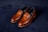 A pair of elegant brown leather loafers with a sleek design and gold detailing on a dark, textured background. The shoes have a polished finish and visible stitching around the edges.