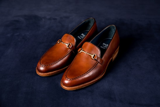 A pair of elegant brown leather loafers with a sleek design and gold detailing on a dark, textured background. The shoes have a polished finish and visible stitching around the edges.