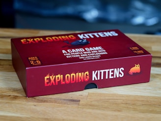 A red box of the card game 'Exploding Kittens' is placed on a wooden surface. The box features bold yellow and white text along with playful graphics, including an image of a cartoon kitten. The game is for ages 7 and up and can be played by 2 to 5 players.