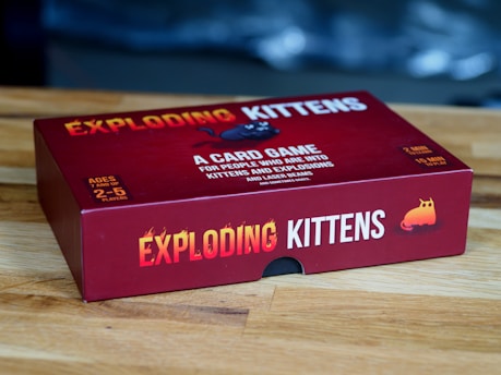 A red box of the card game 'Exploding Kittens' is placed on a wooden surface. The box features bold yellow and white text along with playful graphics, including an image of a cartoon kitten. The game is for ages 7 and up and can be played by 2 to 5 players.