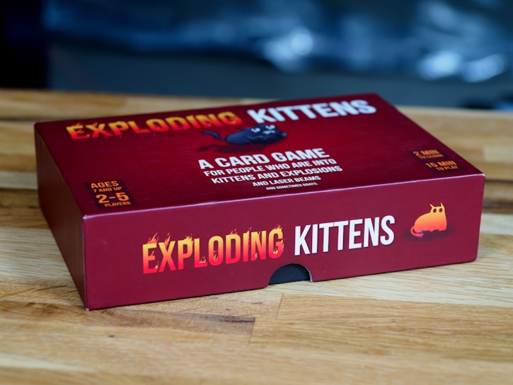 A red box of the card game 'Exploding Kittens' is placed on a wooden surface. The box features bold yellow and white text along with playful graphics, including an image of a cartoon kitten. The game is for ages 7 and up and can be played by 2 to 5 players.