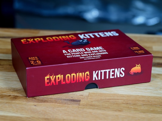 A red box of the card game 'Exploding Kittens' is placed on a wooden surface. The box features bold yellow and white text along with playful graphics, including an image of a cartoon kitten. The game is for ages 7 and up and can be played by 2 to 5 players.