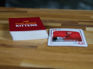 A wooden table features a stack of cards with a red cover labeled 'Exploding Kittens'. Next to it, a single card is laid out showing artwork and text related to the game. The setting is casual and likely related to a tabletop gaming session.